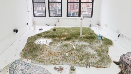 Within a white gallery, an installation sprawls across the ground. The main component is an area where green sprouts grow from a white substance. Additional small components are placed throughout the room.