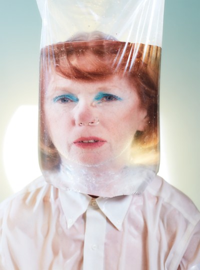 Older woman with bright blue eyeshadow and red hair, seen through a bag full of water. There are bubbles coming out of her nose.