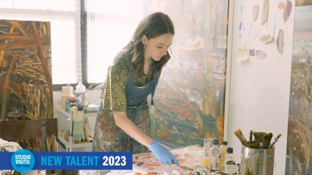 A young woman in a patterned shirt and paint-spattered apron wearing blue gloves holds a palette knife and leans over a table covered with tubes of paint
