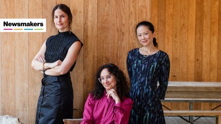 Three women, one seated, in front of a rustic yet refined wood panel wall.