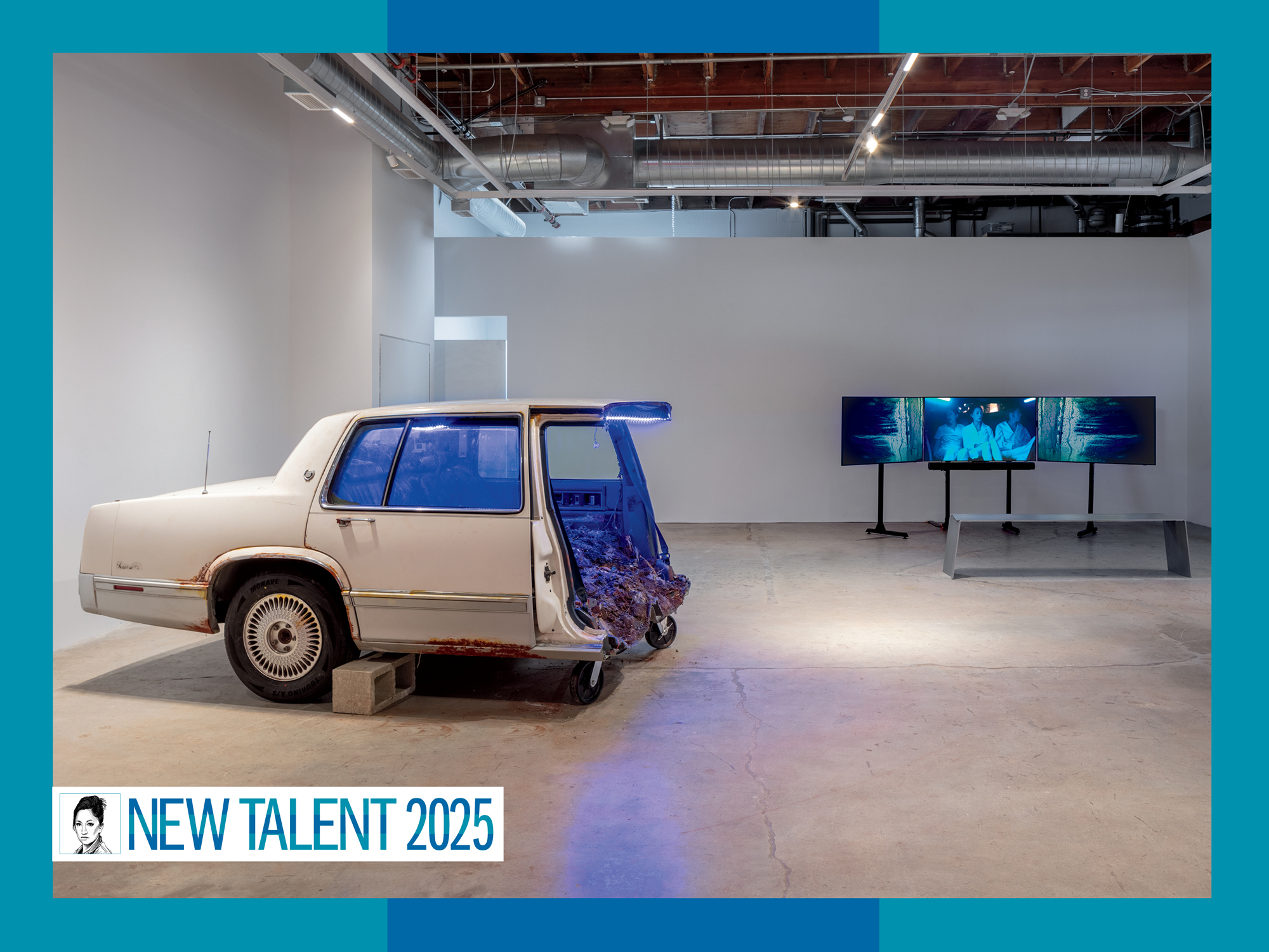In a gallery, an old white car cut in half emits a bleu light, and three screens play a video in the background.