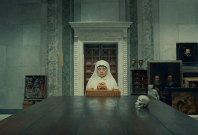 A nun praying at a table. Behind her are paintings leaned against a stone wall.