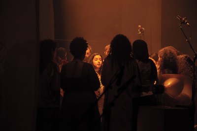 A group of people, with one face appearing to sing or speak in the center.