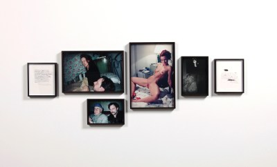 6 framed photos on a wall. Two are of illegible text documents. One is a dark blurry photo of two people having sex. Another is of a woman naked save for red heels, her legs spread. In a third, we see that same woman clothed casually and sitting in bed next to a younger man. Beneath that, that same younger mustachioed man is next to a much older man.