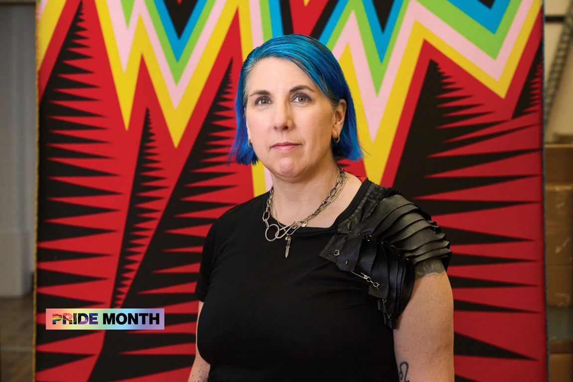 A woman with dyed-blue hair standing in front of a weaving featuring zigzagging lines in various colors.