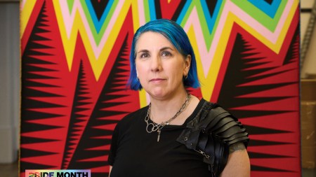 A woman with dyed-blue hair standing in front of a weaving featuring zigzagging lines in various colors.