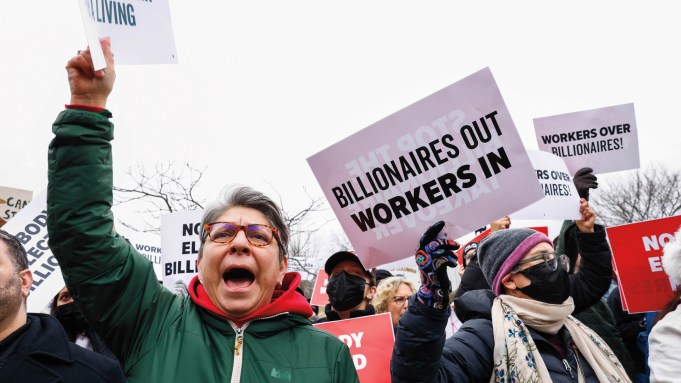 Protestors hold signs and wear jackets and masks. The most visible sign says "Billionaires Out Workers In."