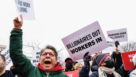 Protestors hold signs and wear jackets and masks. The most visible sign says "Billionaires Out Workers In."