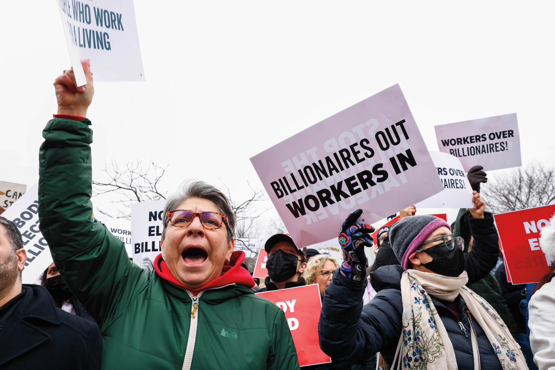 Protestors hold signs and wear jackets and masks. The most visible sign says "Billionaires Out Workers In."