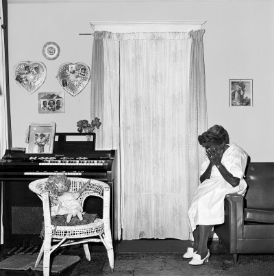A Black woman seated on the arm of a couch with her face in her hands. Hung on the walls of the living room in which she is set are two heart-shaped pictures.