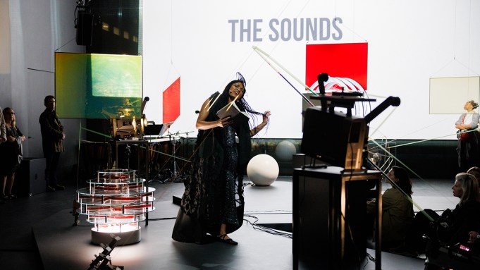 A room is filled with sculptures made of film projectors, as well has colorful squres that dangle in the space. A Black woman with long hair holds a book and wears a long dress as she appears to belt a song.