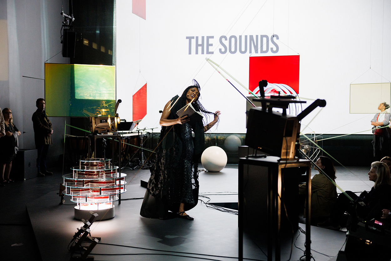 A room is filled with sculptures made of film projectors, as well has colorful squres that dangle in the space. A Black woman with long hair holds a book and wears a long dress as she appears to belt a song.