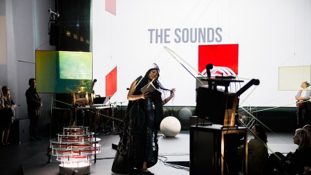 A room is filled with sculptures made of film projectors, as well has colorful squres that dangle in the space. A Black woman with long hair holds a book and wears a long dress as she appears to belt a song.