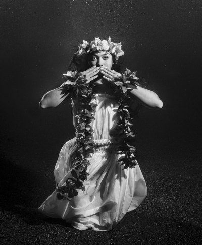 A Native Hawaiian woman wearing white and leis is photographed underwater making a welcome gesture. 