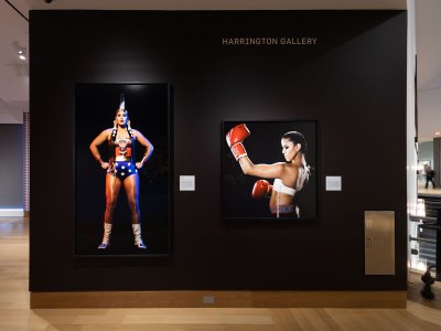 View of a museum wall that is painted black. On it hang two color photographs of Indigenous women, one dressed as Wonder Woman and the other as a boxer.  