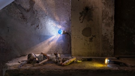 A doll made from reused materials lies on a concrete bench lit by one lightbulb.