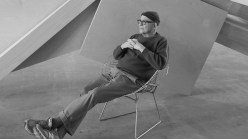 A black-and-white photo of a man in a knit cap with his legs kicked out in a chair in front of a sculpture.