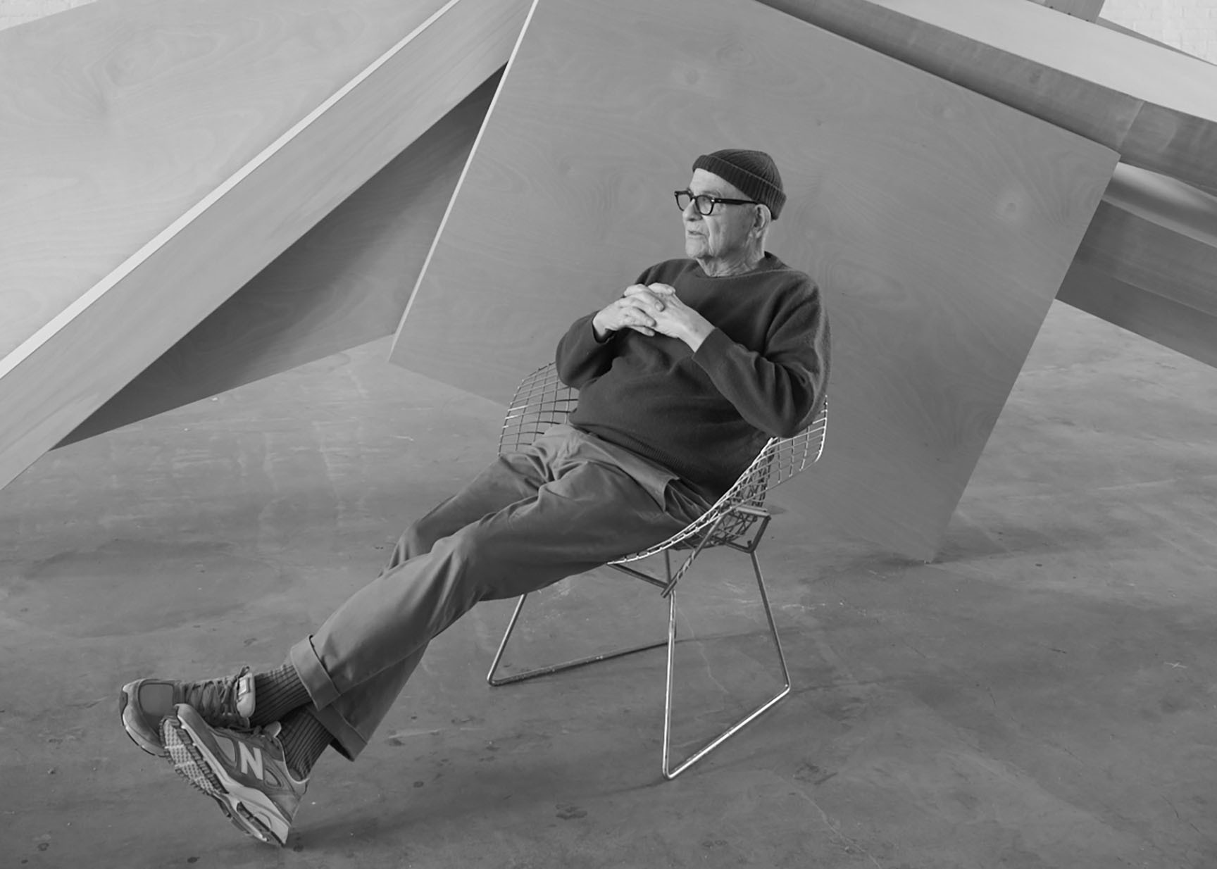 A black-and-white photo of a man in a knit cap with his legs kicked out in a chair in front of a sculpture.