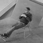 A black-and-white photo of a man in a knit cap with his legs kicked out in a chair in front of a sculpture.