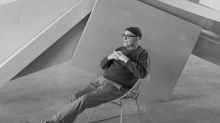 A black-and-white photo of a man in a knit cap with his legs kicked out in a chair in front of a sculpture.