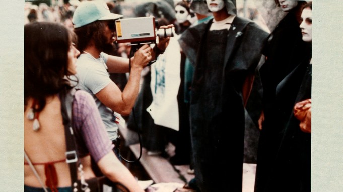 Two people hold vintage video equpimnet at a group of people dressed in all black with their faces painted white.
