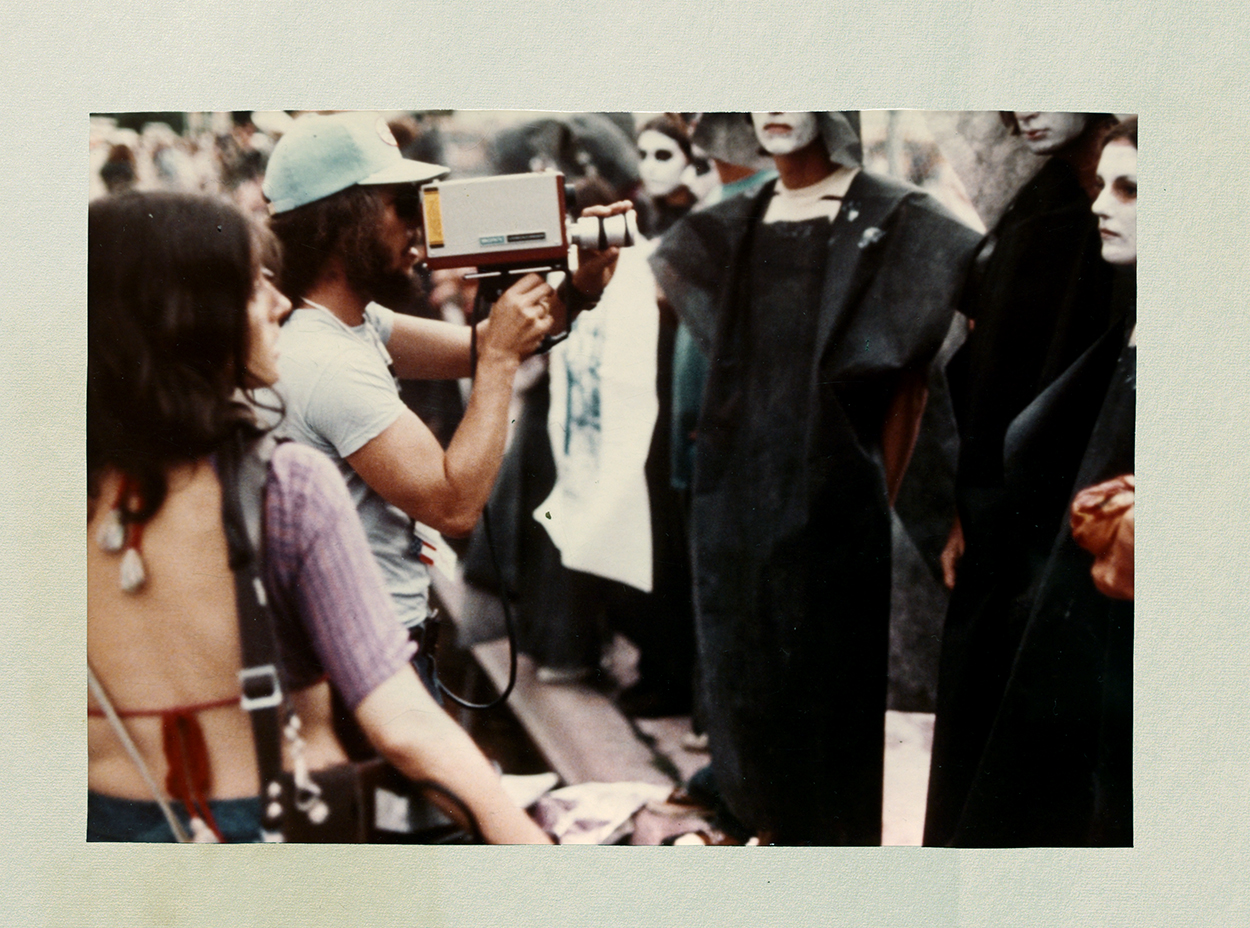 Two people hold vintage video equpimnet at a group of people dressed in all black with their faces painted white.