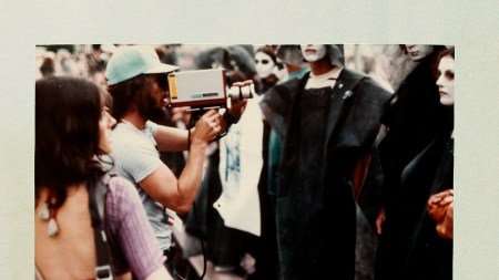 Two people hold vintage video equpimnet at a group of people dressed in all black with their faces painted white.