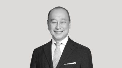 A silhouetted black-and-white portrait of a smiling Asian man in a suit and tie on a gray background.