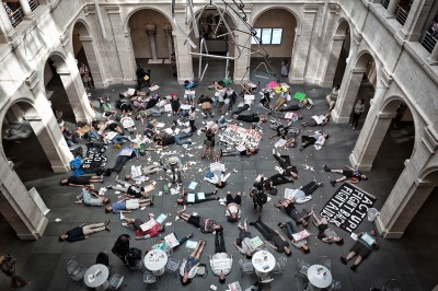 A bunch of bodies lie on the floor of a museum atrium amid pill bottles and protest signs.