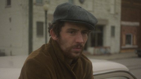 Josh O'Connor in a gray hat and brown jacket leans across a 1970s car.