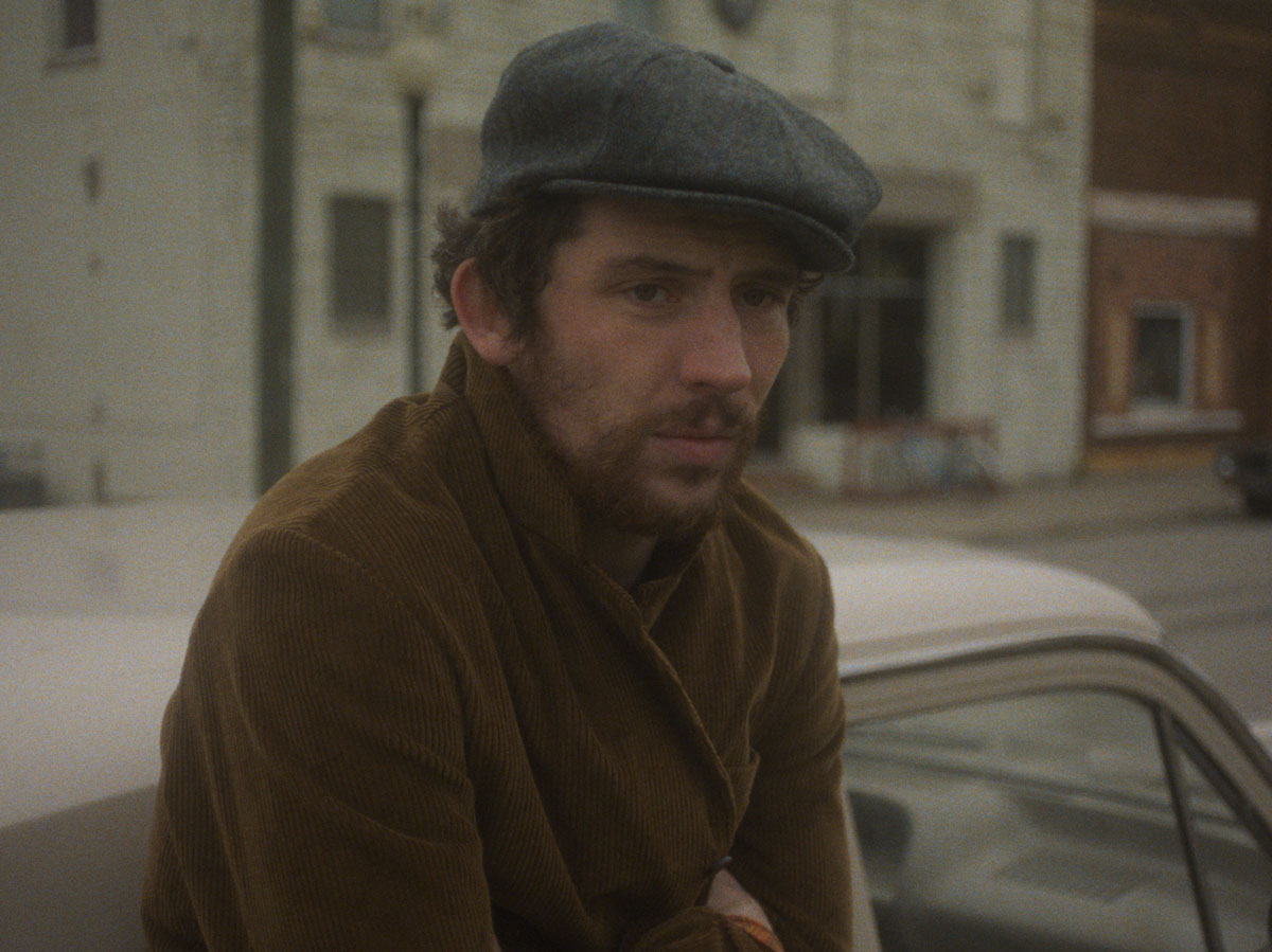 Josh O'Connor in a gray hat and brown jacket leans across a 1970s car.