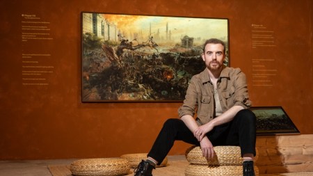 A white man with a goatee and short hair sits in front of a digital painting.