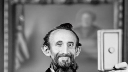 A square vintage photo of a deformed looking Abraham Lincoln holding a rectangular object in front of his face.