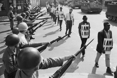Grayscale photo of Black men wearing signs that say I Am A Man as white men with helmets point their long guns at them.