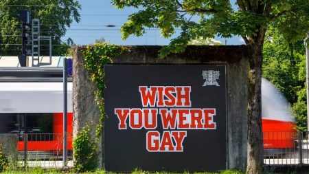 A billboard reading 'WISH YOU WERE GAY' in red lettering against a black background. A train whizzes by behind it.