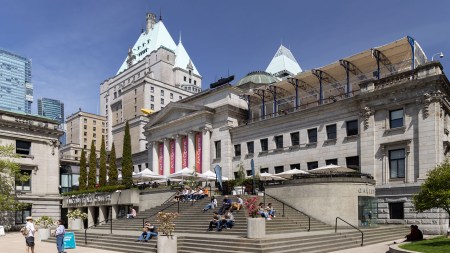 The front exterior of the Vancouver Art Gallery on a sunny day, showing several people sitting on its steps.