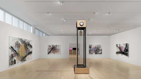 Installation view of a museum exhibition with a large sculpture that looks like a grandfather clock with a piece of granite suspended in its center. On the walls are artworks showing black-and-white details of trees over grids.