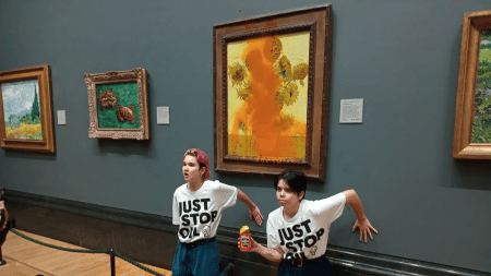 Two white protesters in tee shirts that say "just stop oil" have their hands glued to the wall in front of Van Gogh's sunflowers, which is covered in tomato soup.