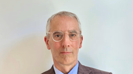 A man with glasses and white hair wearing a brown suit and tie.