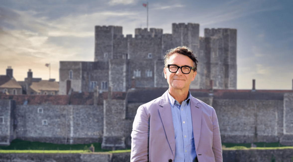 A man in a purple suit and glasses stands in front of a castle.